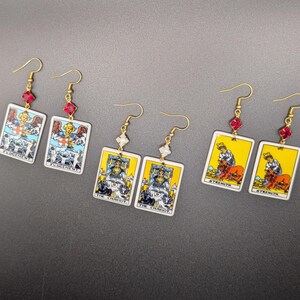 May include: Gold-tone earrings featuring tarot card designs. The earrings display images of "Judgement", "The Chariot", and "Strength" cards. Each earring has a decorative red or clear crystal accent and a gold hook.