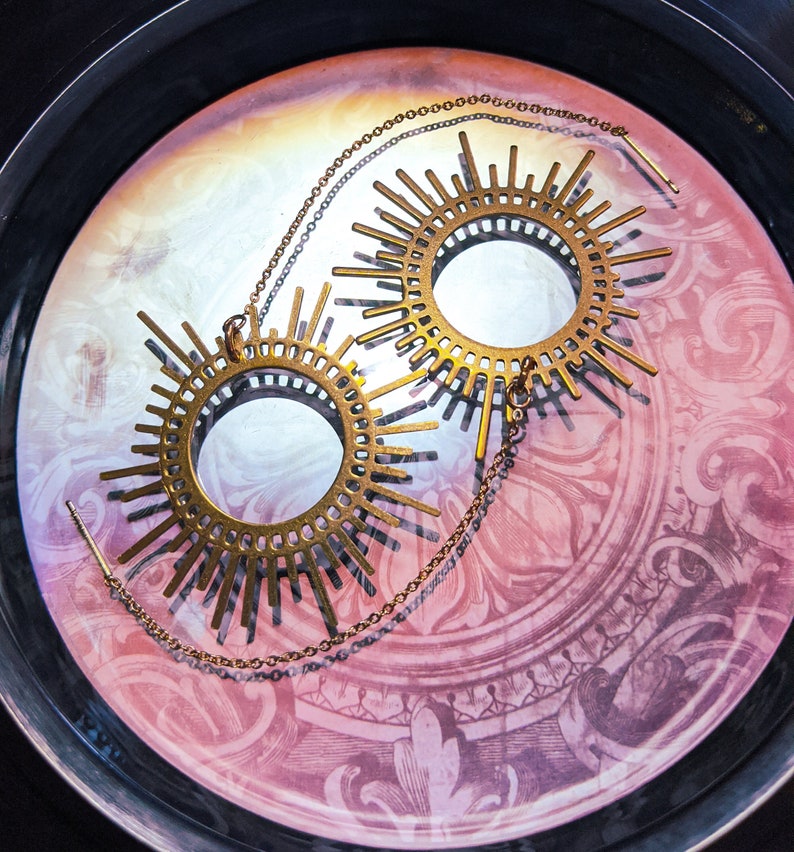 May include: A pair of gold-toned earrings with a sunburst design. Each earring features a circular center with a delicate pattern and multiple rays extending outward.