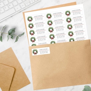 May include: A sheet of rectangular white address labels featuring a watercolor illustration of a green Christmas wreath with a red bow. Each label includes the placeholder text NAME SURNAME, 741 Old State Road, and Pine View, IL 33103.
