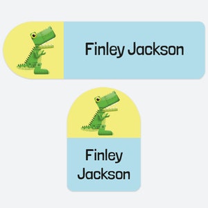 May include: Two rectangular adhesive labels with rounded corners featuring a green mechanical dinosaur illustration on a yellow background and the name Finley Jackson printed in black text on a light blue background for identification purposes.
