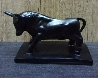 Pode incluir: Uma figura de touro preto com a cauda levantada, em pé sobre uma base preta. O touro é feito de um material liso e polido.