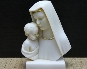 May include: A white marble bust sculpture of a mother and child. The mother wears a draped head covering and a gold necklace. The child is wrapped in a cloth. The sculpture is mounted on a white rectangular base.
