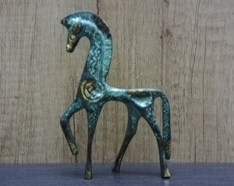 Puede incluir: Figura de caballo de color bronce con un diseño estilizado y delgado. El caballo tiene un cuello y patas alargadas, con un remolino decorativo en su costado. La figura está sobre un fondo de madera.