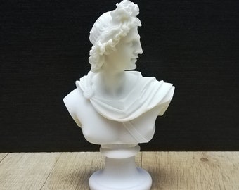 May include: White plaster bust sculpture of a man with a laurel wreath on his head. The bust is on a white pedestal.