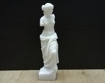 Op de afbeelding: Witte gipsen beeld van een staande vrouw met een gedrapeerde doek. Het beeld staat op een houten oppervlak.