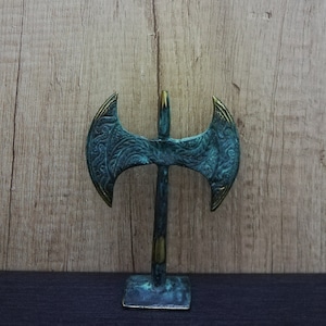 May include: A decorative double-headed axe sculpture with a green patina finish and gold accents. The axe is mounted on a small base and has intricate details on the blades.