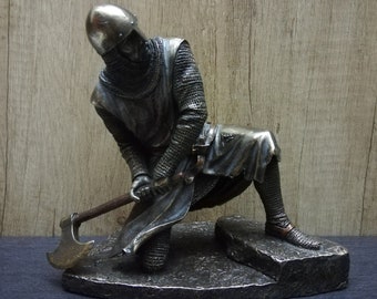 May include: A silver knight figurine kneeling with a battle axe in hand. The knight is wearing chainmail armor and a helmet. The figurine is on a rocky base.