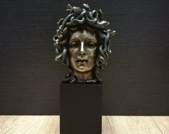 Puede incluir: Una escultura en bronce de la cabeza de Medusa, con serpientes detalladas en lugar de cabello. La escultura está montada sobre un pedestal rectangular negro, que representa la figura mitológica griega.