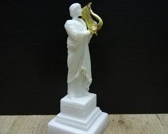 Op de afbeelding: Wit marmeren beeld van een vrouw die een gouden lier bespeelt. Het beeld staat op een witte marmeren voet.