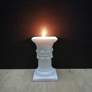 Peut inclure: Un bougeoir blanc en forme de colonne miniature avec des détails décoratifs. La bougie est allumée et a une flamme qui brûle à l'intérieur.