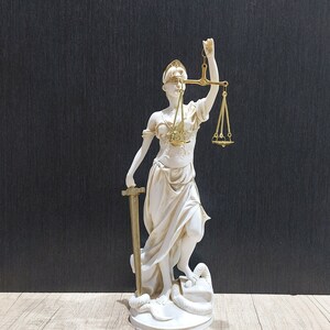 Puede incluir: Una estatua blanca de la figura de la Justicia, con los ojos vendados, sosteniendo una espada y una balanza. La estatua está de pie sobre una base con una serpiente enroscada alrededor.