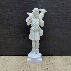 May include: White marble statue of a shepherd carrying a lamb on their shoulder. The shepherd is wearing a tunic and sandals. The statue is standing on a square base.