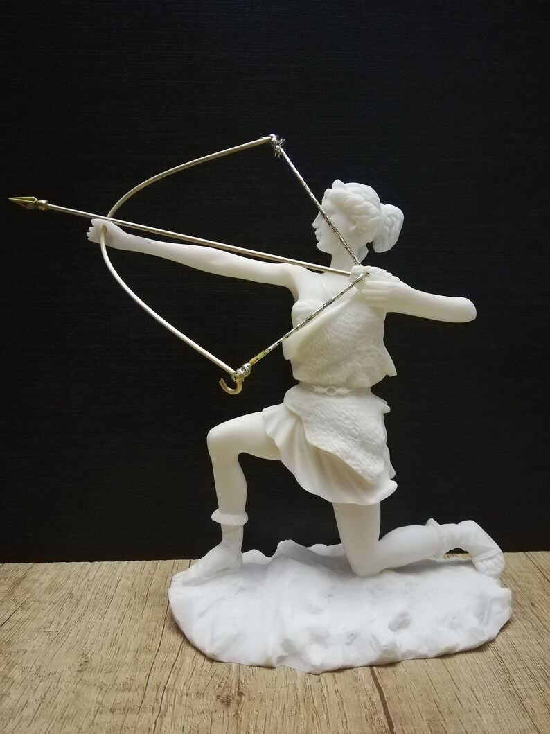 May include: White marble statue of a woman kneeling and holding a bow and arrow. The statue is on a white marble base.