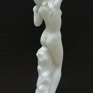 May include: White marble statue of a woman standing on a square base. The woman is nude and has her arms raised above her head. The statue is about 10 inches tall.