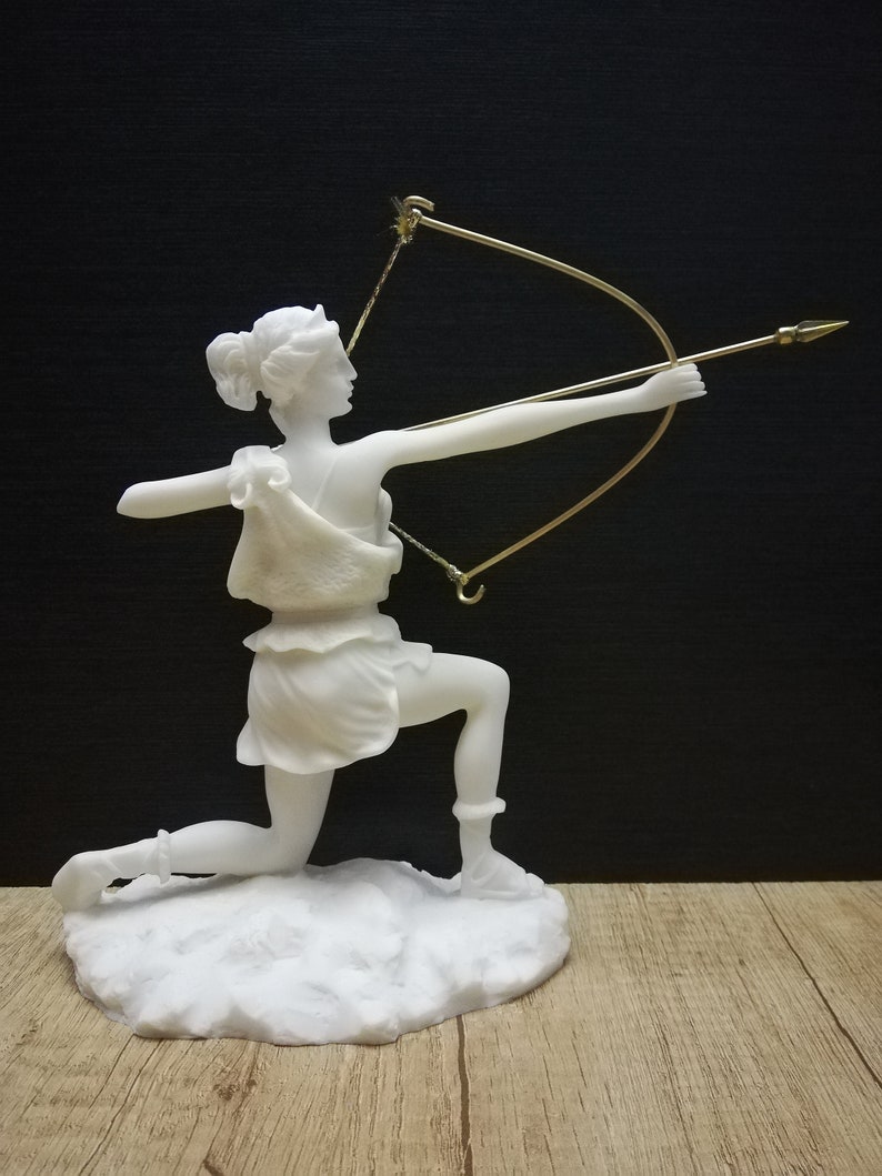 May include: White marble statue of a woman kneeling and holding a bow and arrow. The statue is on a white marble base.