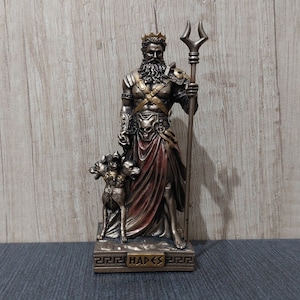 Peut inclure: Statue détaillée d'Hadès, dieu grec des enfers, tenant un trident et accompagné de Cerbère. La statue est principalement en bronze et or, avec une robe rouge et bronze. La base de la statue indique "HADES".