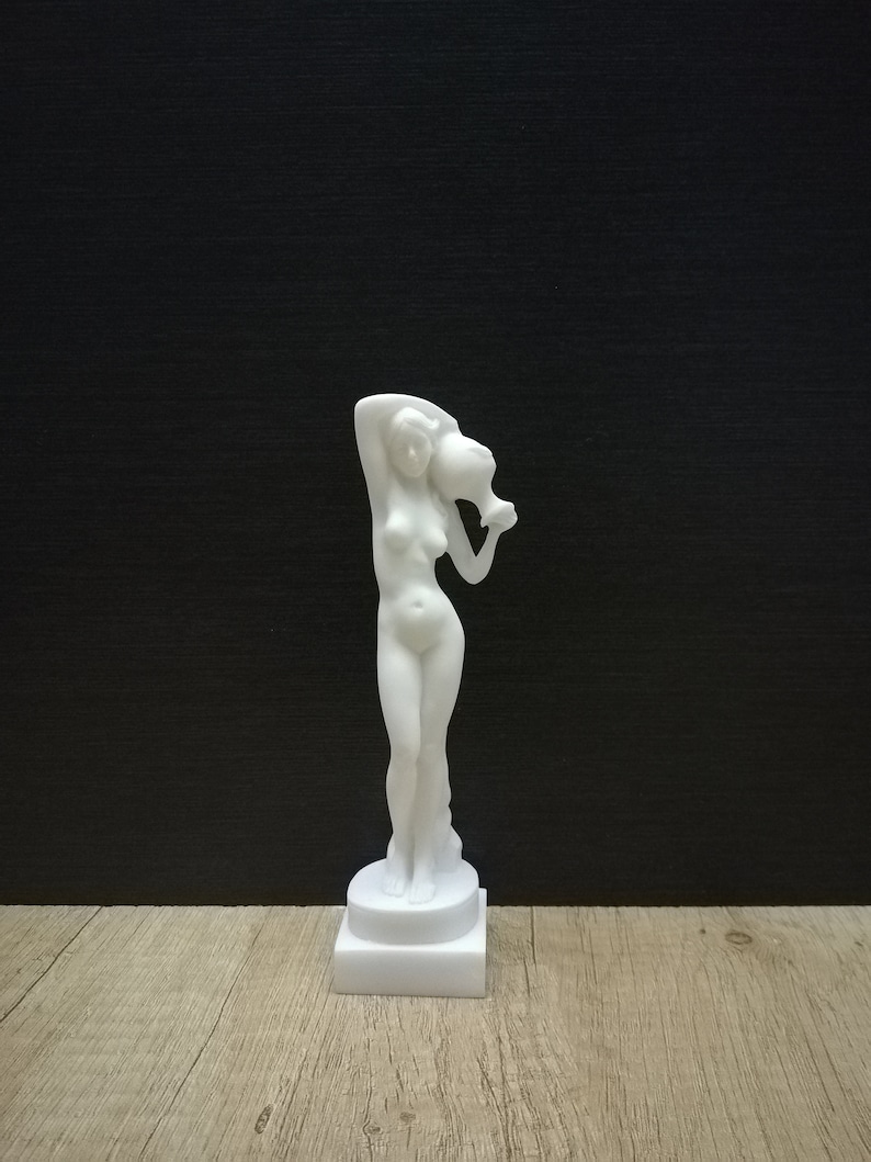 May include: White marble statue of a woman holding a pitcher above her head. The statue is standing on a square base.