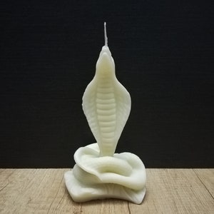 May include: White candle shaped like a cobra with its head raised and a coiled body. The candle is on a wooden surface.