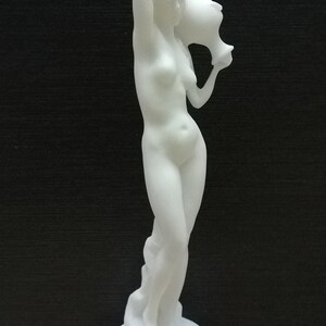 May include: White alabaster sculpture of a woman holding a pitcher. The sculpture is on a white base.
