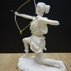 May include: White marble statue of a woman kneeling and holding a bow and arrow. The statue is on a white marble base.