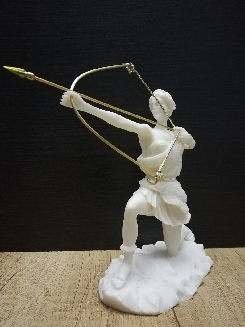 May include: White marble statue of a figure holding a bow and arrow, the figure is kneeling and looking to the right. The statue is on a white marble base.