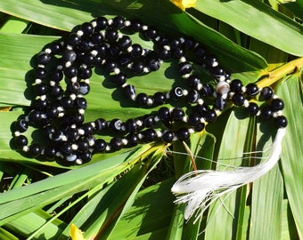 May include: Black and white agate prayer beads with a white tassel, resting on green leaves.