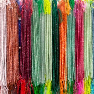May include: A variety of colorful gemstone bead strands, including green, red, orange, and white, are displayed for sale. The strands are hung on a wall and are ready to be purchased.