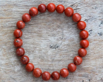 May include: A red jasper beaded bracelet with a circular shape. The bracelet is made of smooth, round beads and is laid out on a wooden surface.