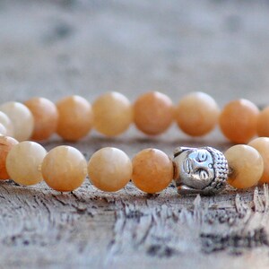 Puede incluir: Un brazalete de piedras preciosas de color naranja claro con un dije de Buda plateado. El brazalete est&aacute; hecho de cuentas redondas y est&aacute; enhebrado en un cord&oacute;n el&aacute;stico.