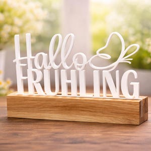 Op de afbeelding: Een wit decoratief bord met de tekst "Hallo Frühling" en een vlinderontwerp. Het bord is gemonteerd op een rechthoekige houten basis. De achtergrond toont vage bloemen en natuurlijk licht.