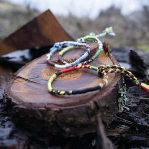 Könnte beinhalten: Eine Sammlung farbenfroher Freundschaftsarmbänder liegt auf einer verwitterten Holzoberfläche. Die Armbänder weisen verschiedene Muster und Farben auf, darunter Blau, Grün, Rot, Gelb und Schwarz. Der Hintergrund ist unscharf, was auf eine Umgebung im Freien hindeutet.