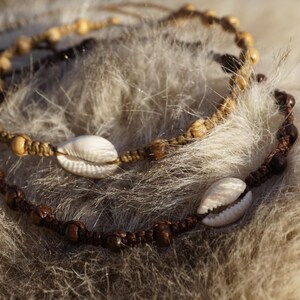 Könnte beinhalten: Zwei handgefertigte Armbänder mit natürlichen Elementen. Ein Armband ist hellbraun mit Holzperlen und einer weißen Muschel. Das andere ist dunkelbraun mit Holzperlen und einer weißen Muschel. Die Armbänder liegen auf einer flauschigen, hellbraunen Oberfläche.