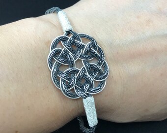 May include: A silver bracelet with a black braided design and a large, intricate knot in the center.