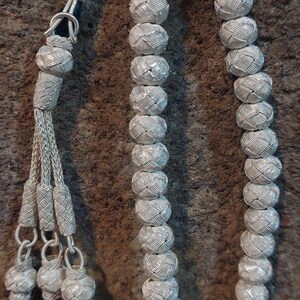May include: A silver prayer beads necklace with a braided tassel. The beads are round and have a woven pattern. The tassel is made of silver wire and has a blue bead at the end.