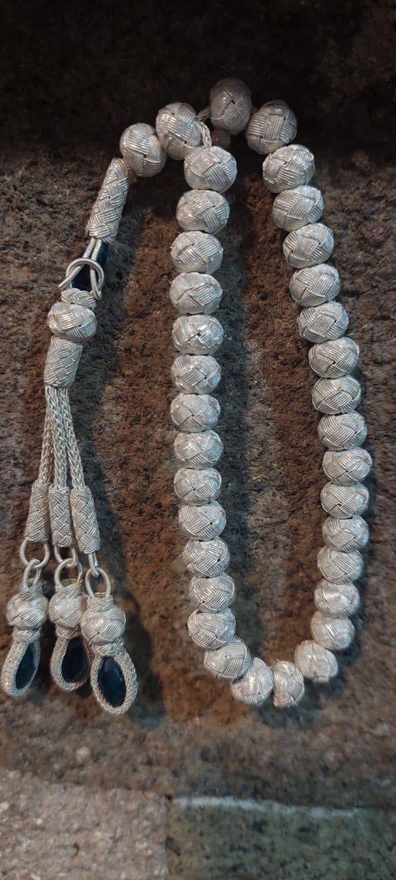 May include: A silver prayer beads necklace with a braided tassel. The beads are round and have a woven pattern. The tassel is made of silver wire and has a blue bead at the end.