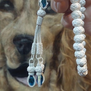 May include: A silver prayer beads necklace with blue gemstone accents. The beads are woven in a tight pattern and the tassel has three strands with blue gemstones at the end.