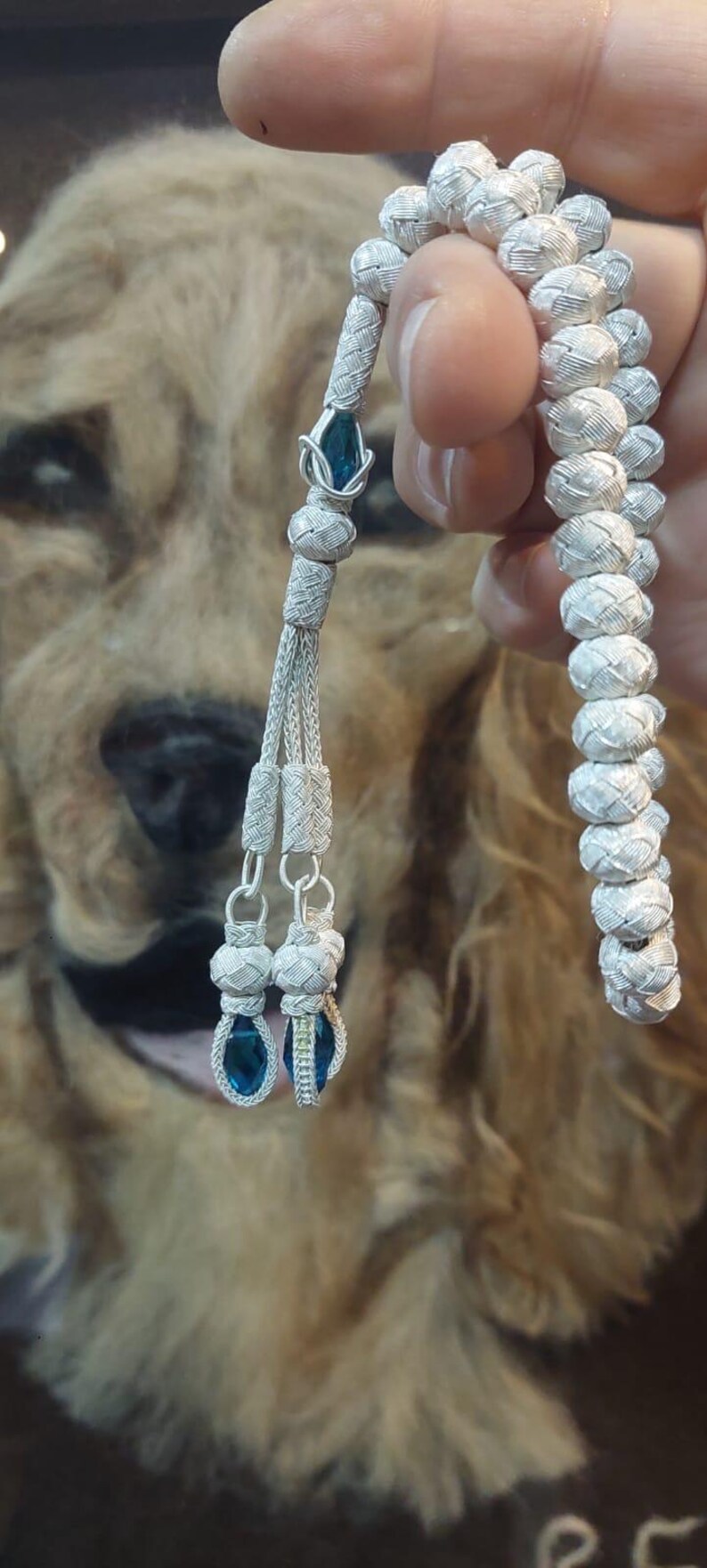 May include: A silver prayer beads necklace with blue gemstone accents. The beads are woven in a tight pattern and the tassel has three strands with blue gemstones at the end.