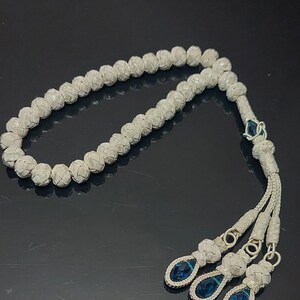 May include: A silver prayer beads necklace with blue gemstone accents. The beads are woven together in a knot pattern.
