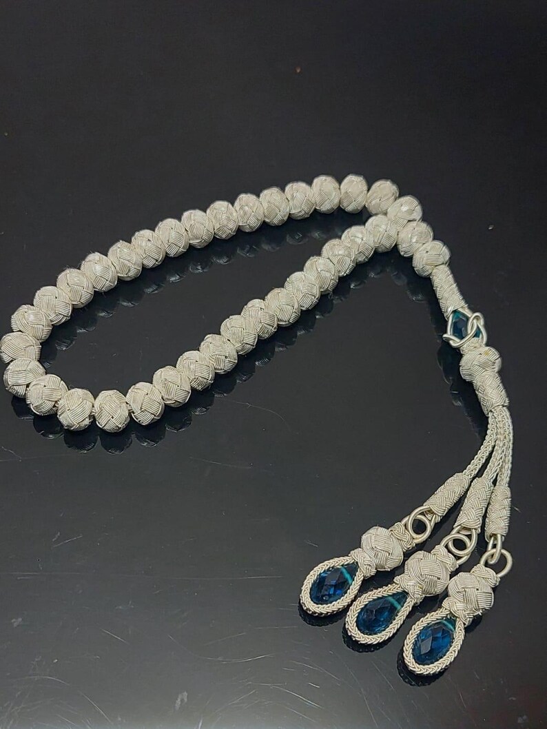 May include: A silver prayer beads necklace with blue gemstone accents. The beads are woven together in a knot pattern.