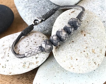 May include: A silver bracelet with a braided design and multiple woven beads. The bracelet is photographed on a wooden surface with several gray stones.
