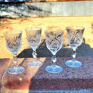 May include: Four clear crystal wine glasses with intricate starburst and floral patterns. Each glass has a faceted stem and a round base. The glasses are arranged on a brick surface, with sunlight casting shadows.