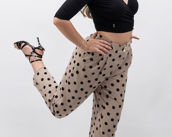 May include: A woman wearing a black top and beige polka dot pants. She is wearing black heels and has her leg raised.