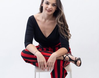 May include: A woman wearing a black top and red and black striped pants sits on a white stool. She is wearing black heeled sandals.