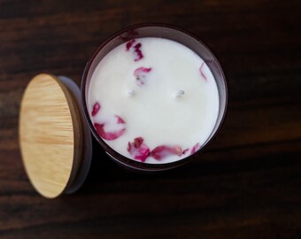 May include: A cylindrical candle in a dark glass container with a white wax surface embedded with pink rose petals and two wicks, accompanied by a circular bamboo lid placed to the side of the candle vessel.