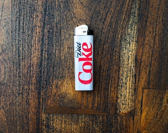 May include: A white lighter with the words "Diet Coke" in red script. The lighter is rectangular and has a metal top. The background is a dark wood surface.