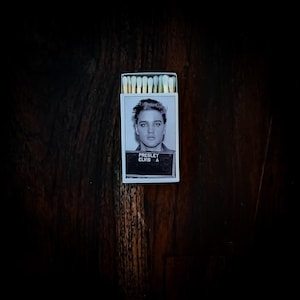 Puede incluir: Una caja de cerillas con una imagen en blanco y negro de la foto policial de Elvis Presley. La caja tiene un borde blanco y el texto "PRESLEY ELVIS A" es visible. Las cerillas tienen puntas de color amarillo claro y están bien dispuestas.