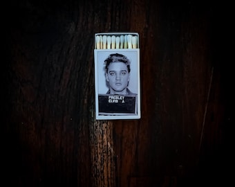 May include: A matchbox featuring a black and white image of Elvis Presley's mugshot. The matchbox has a white border and the text "PRESLEY ELVIS A" is visible. The matches have light yellow tips and are arranged neatly.