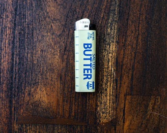 May include: A novelty lighter with a cream-colored body and the word "BUTTER" printed in blue. The lighter also has the words "SALTED" and "AA" printed on it. The lighter is on a dark brown wooden surface.