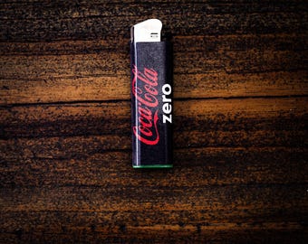 May include: A black rectangular lighter featuring the red Coca-Cola logo and the word zero printed in white text on the front. The lighter has a metallic silver ignition mechanism at the top and a green base at the bottom.