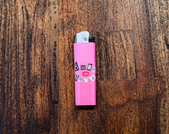 May include: A pink lighter with the words "Burn Book" in a collage style. The lighter has a silver metal top and is set against a dark wood grain background. The text includes a red lip graphic.